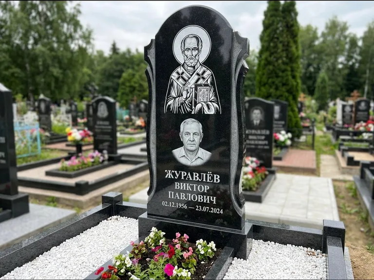 Памятник с гравировкой иконы: стела с образом в полный рост на обороте — гравировка образа Матроны Московской, низкие кусты спиреи в цвету, работа «Лебедева» в Воронеже
